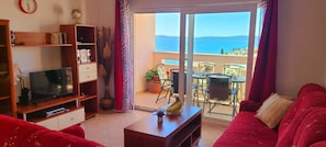 Una sala de estar con televisión, estantería, sofá rojo y balcón con mesa y sillas.