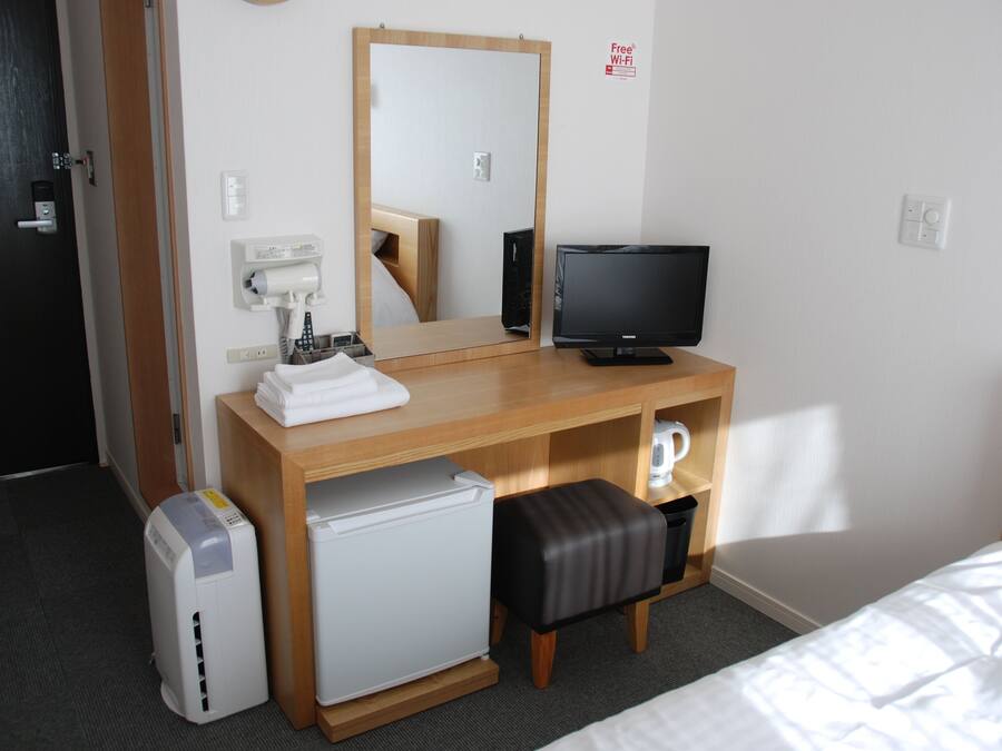 A hotel room with a wooden desk, a mirror, a television, a mini-fridge, a coffee machine, and a small seating area.