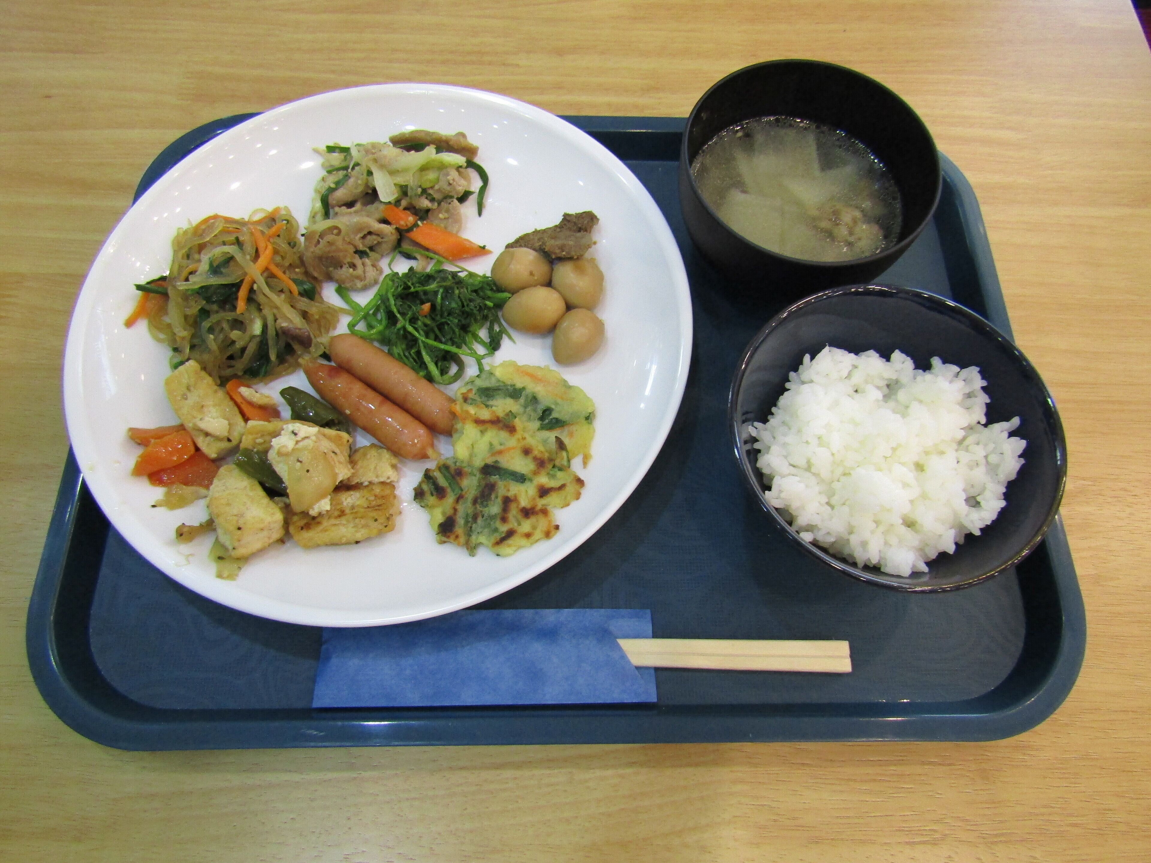 A tray with a plate of food including vegetables, tofu, and sausages, a bowl of rice, and a bowl of soup.