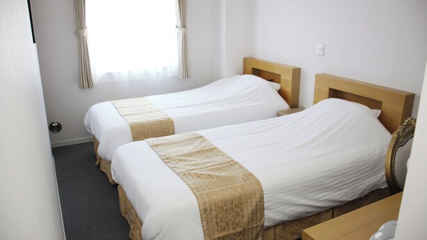 A hotel room with two single beds, a wooden headboard, a small wooden desk, and a window with curtains. - Business Hotel Yamate Inn (Kofu)