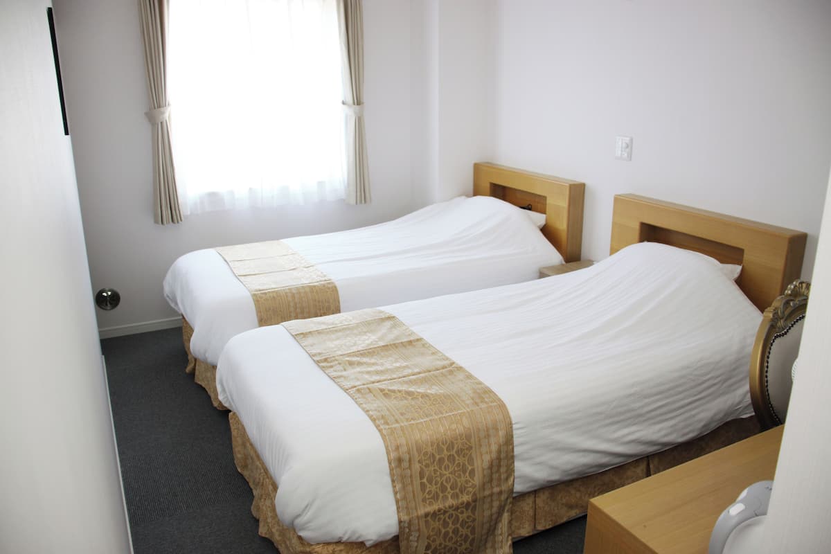A hotel room with two single beds, a wooden headboard, a small wooden desk, and a window with curtains.