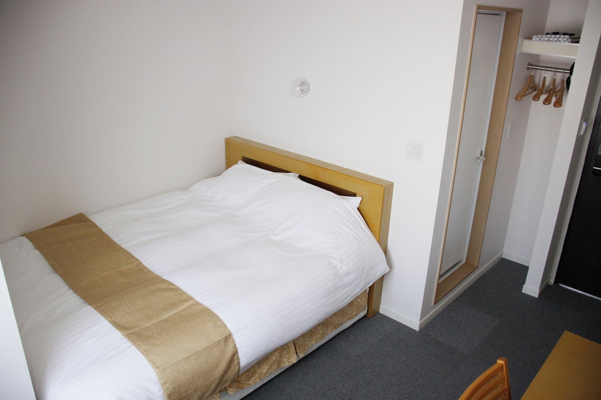 A single bed with a wooden headboard, a white pillow, and a beige blanket. There is a door to the right with a coat rack above it.