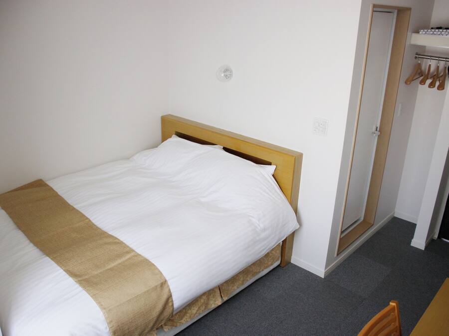 A single bed with a wooden headboard, a white pillow, and a beige blanket. There is a door to the right with a coat rack above it.