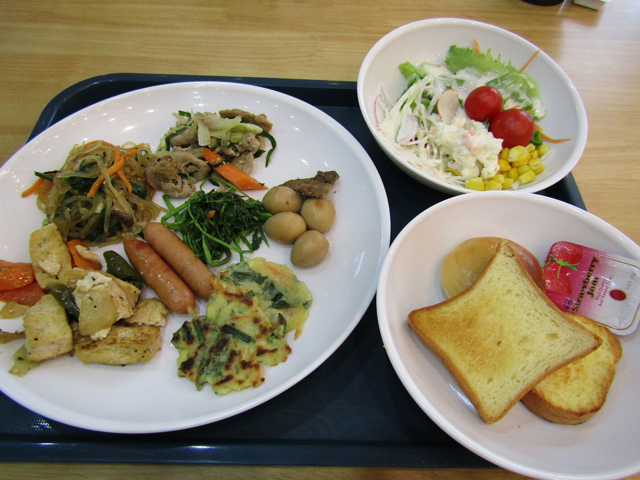 Una bandeja con un plato de comida que incluye salchichas, verduras y una ensalada, junto a otro plato con tostadas y mermelada.
