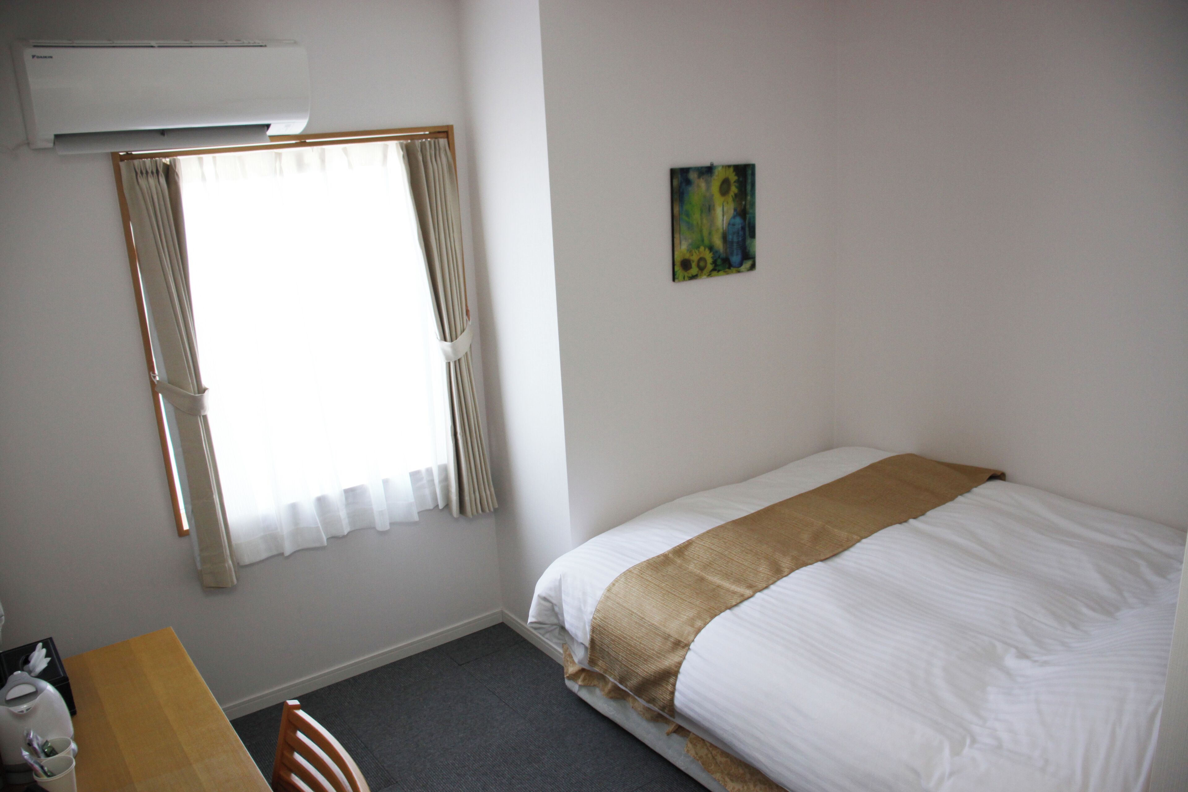 A hotel room with a bed, a table, a chair, a window with curtains, an air conditioner, and a painting on the wall.