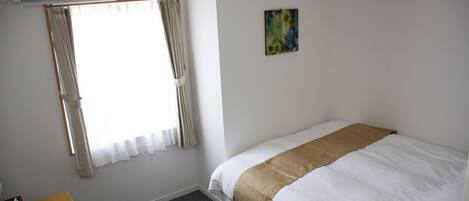 A hotel room with a bed, a table, a chair, a window with curtains, an air conditioner, and a painting on the wall.