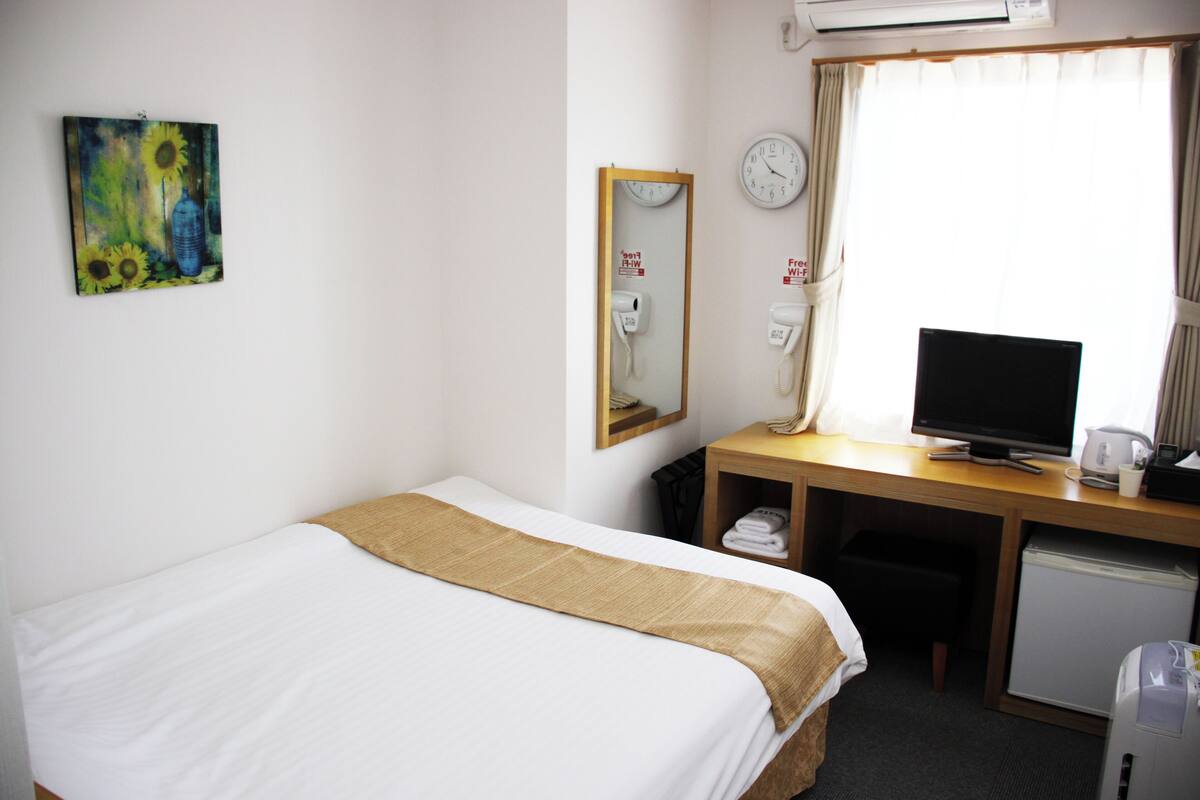A hotel room with a bed, a desk with a computer, a TV, an air conditioner, and a window with curtains.