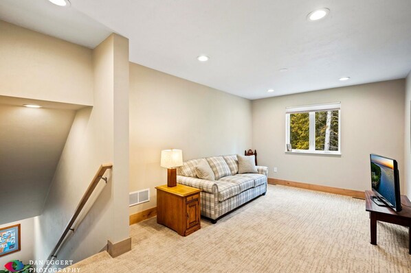 A spacious room with a plaid sofa, a wooden nightstand with a lamp, a TV on a stand, and a window with a view of trees.