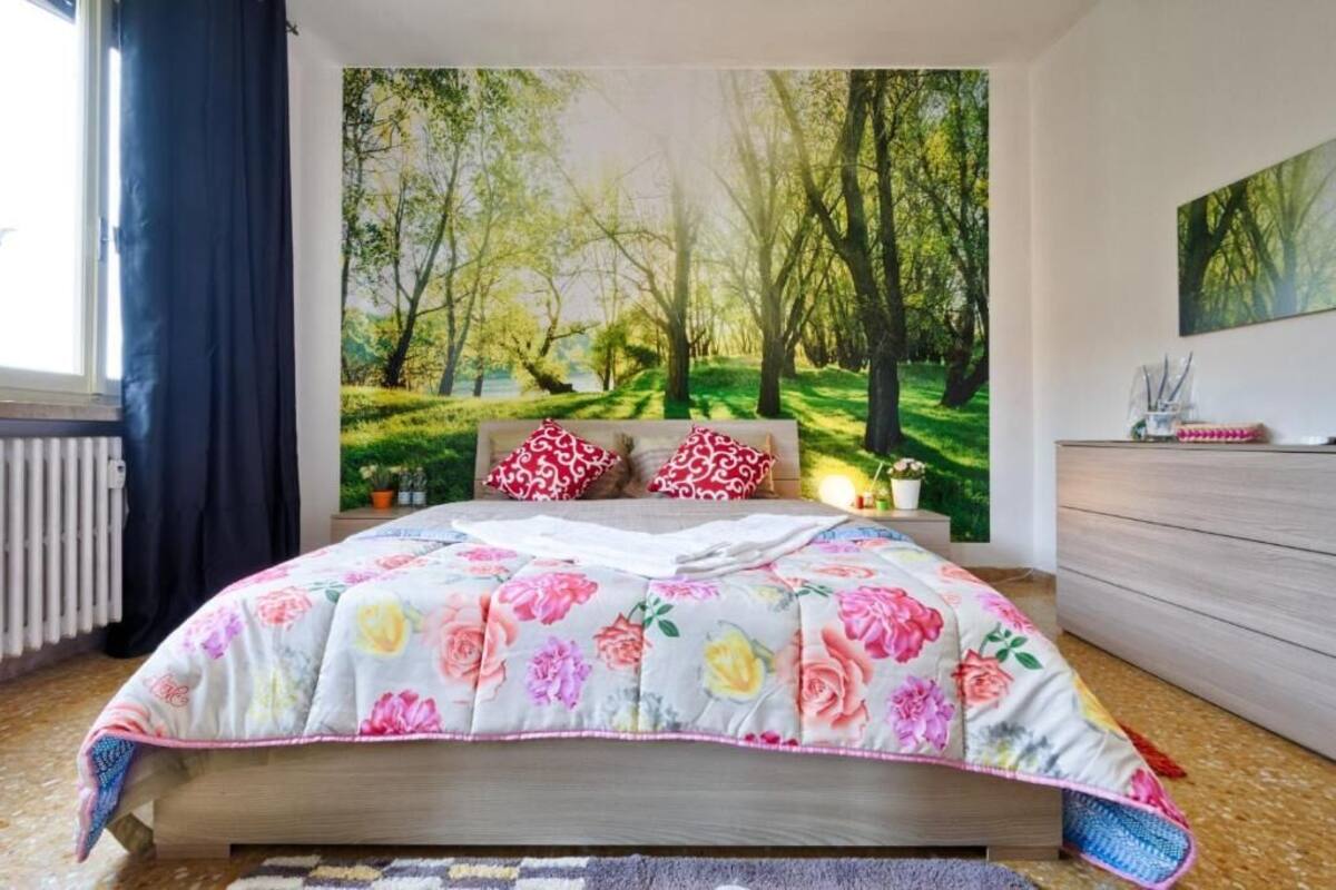 A bedroom with a floral bedspread, a view of a forest through a window, and a wooden dresser.