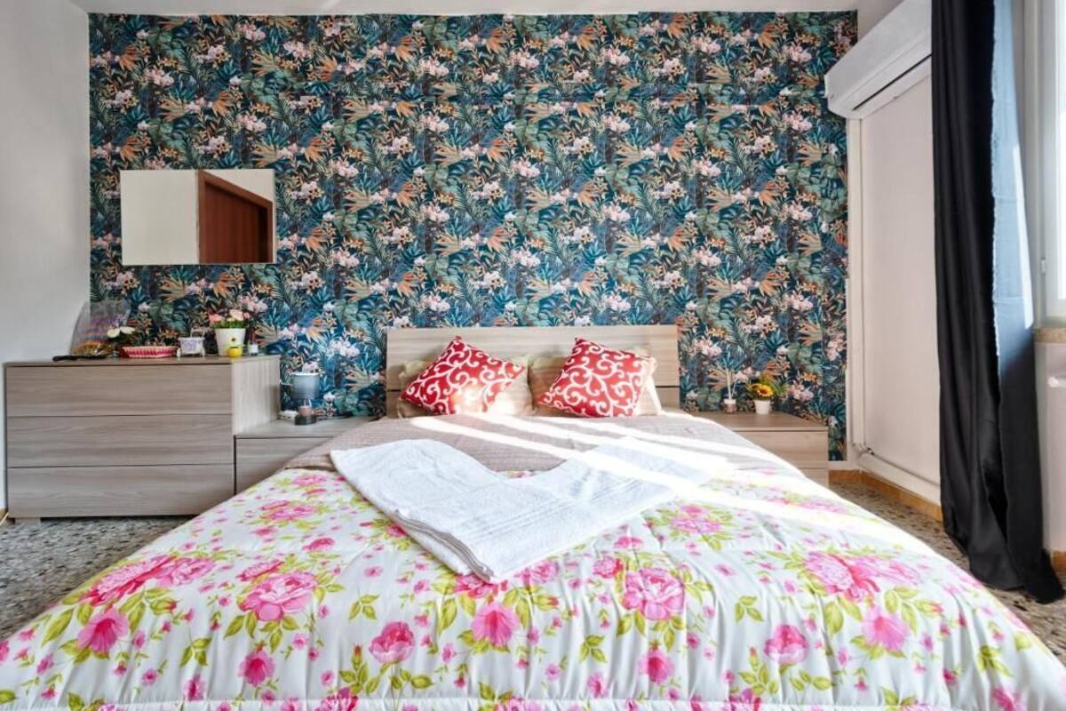 A bedroom with a floral wallpaper, a bed with a floral bedspread, two nightstands with decorative items, and a mirror on the wall.
