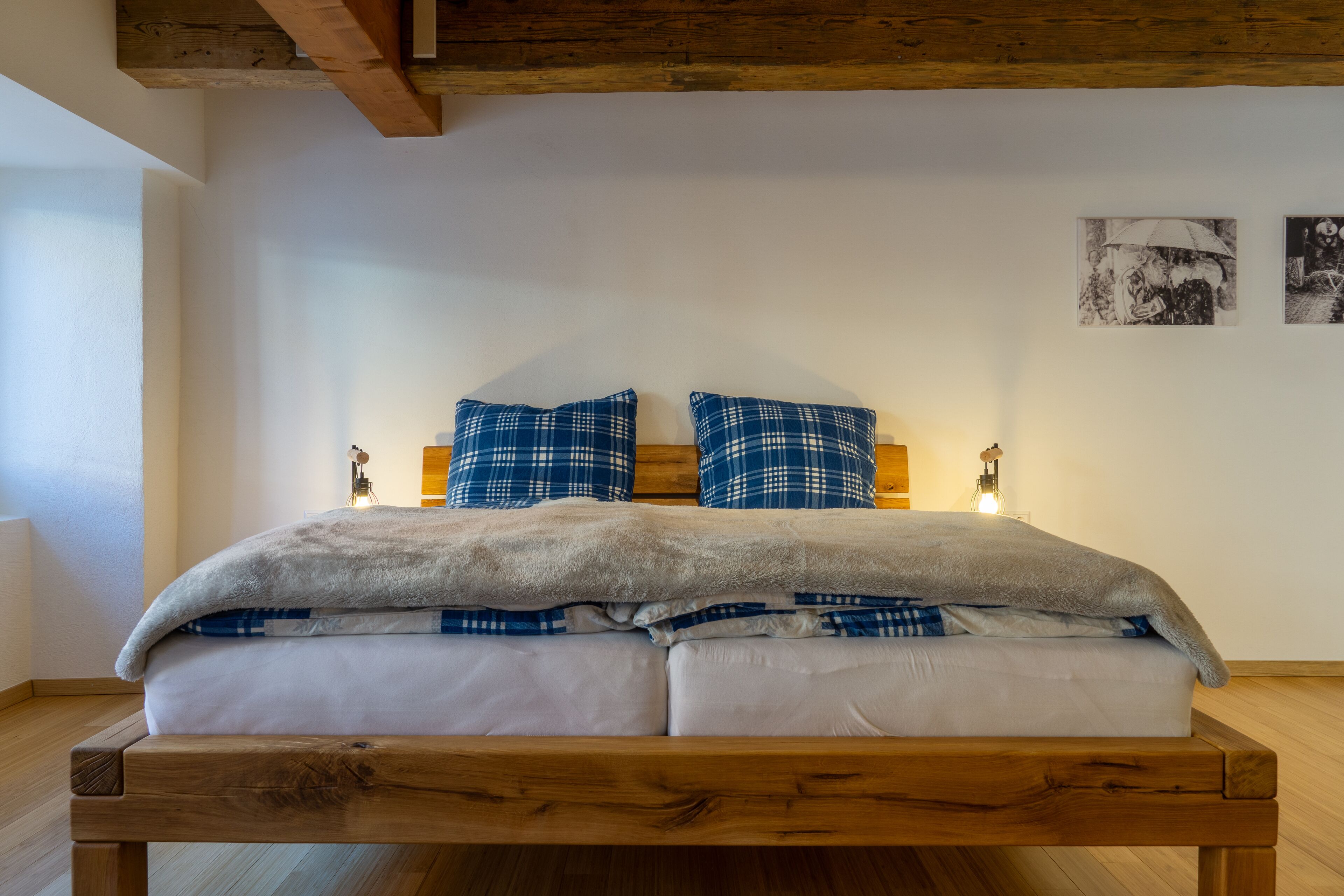 A bed with a wooden frame, white bedding, and blue plaid pillows. There are two wall-mounted lights on either side of the bed.