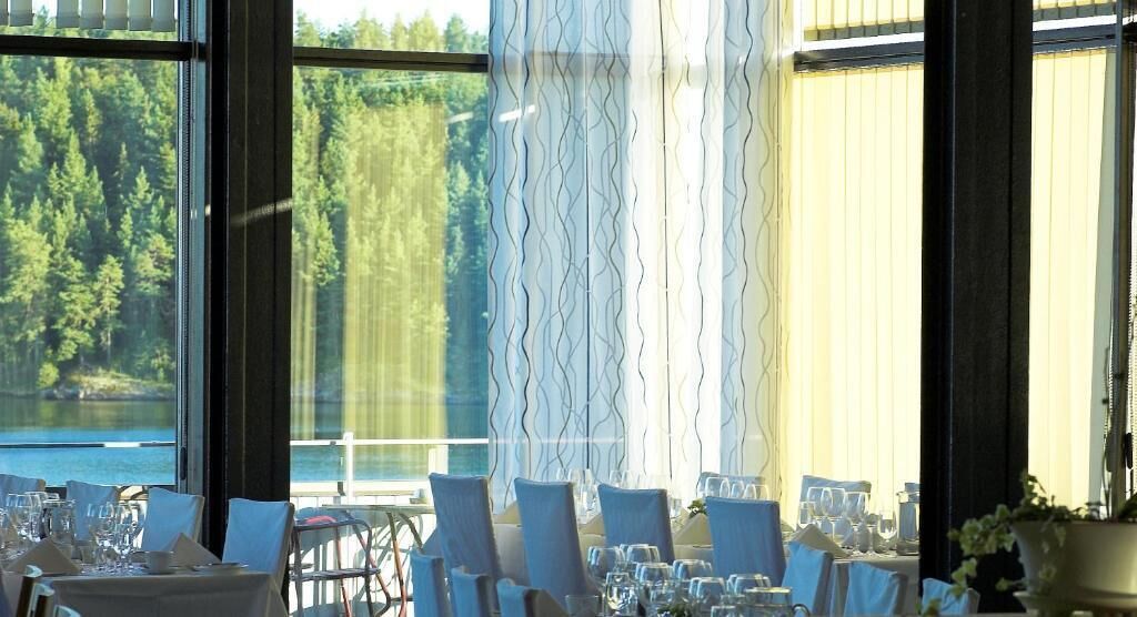 Un restaurante con mesas dispuestas para comer, amplias ventanas con vistas a un bosque y un lago.
