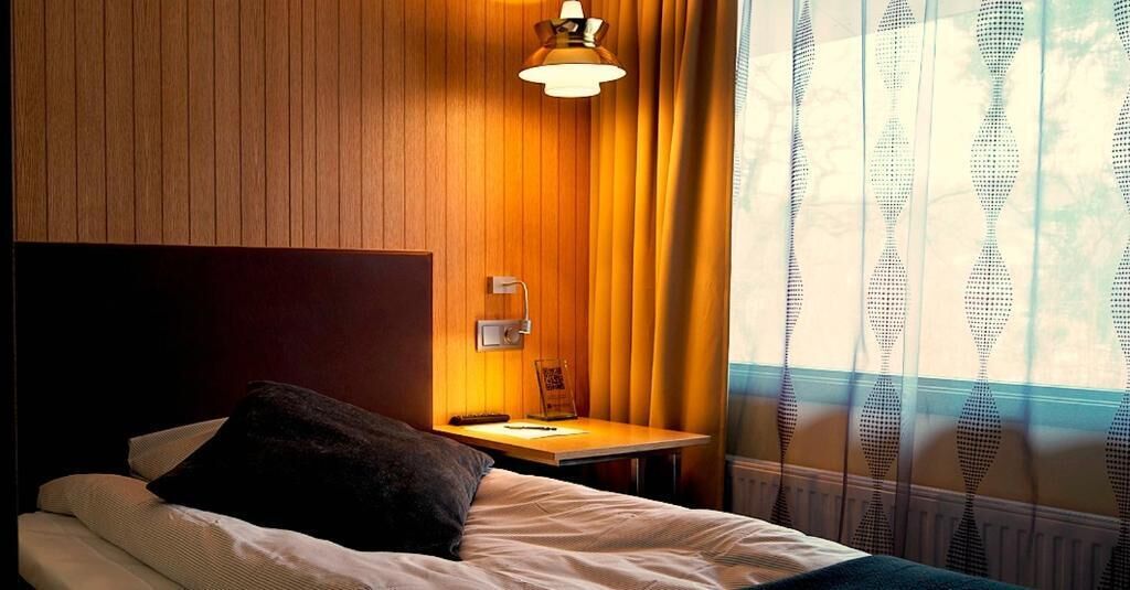 Habitación de hotel con pared de paneles de madera, una mesita de noche con lámpara, una cama con almohada y una ventana con cortinas estampadas.