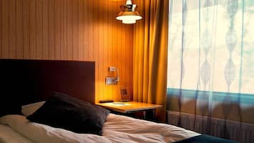 Habitación de hotel con pared de paneles de madera, una mesita de noche con lámpara, una cama con almohada y una ventana con cortinas estampadas.