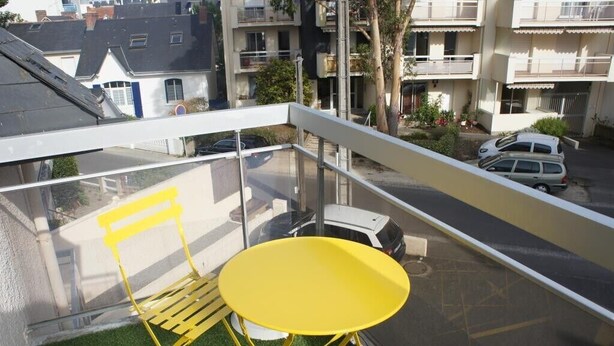 Un balcon avec une chaise et une table jaunes, donnant sur une rue bordée de voitures garées et d’immeubles d’habitation.