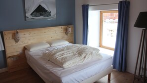 A bedroom with a wooden headboard, a bed with white bedding, a wall-mounted lamp, and a window with blue curtains. - Chalet vue Imprenable (Montvalezan)