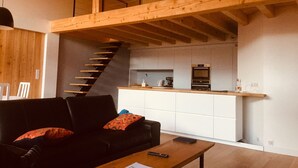 A modern loft-style living space with a wooden staircase, a kitchen area, and a black leather sofa with red cushions. - Chalet vue Imprenable (Montvalezan)