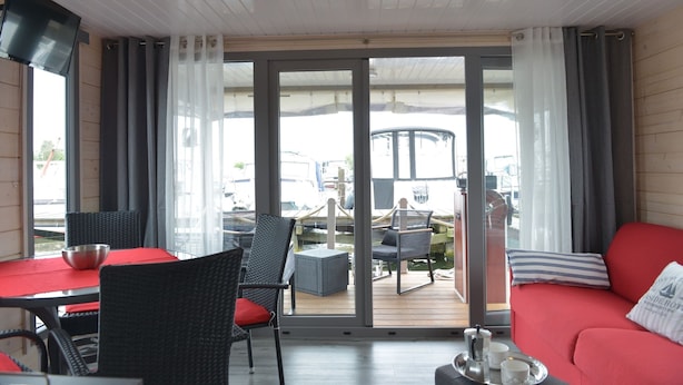 A room with a red sofa, a dining table with chairs, a TV, and a view of boats through large glass doors. - Time out on the Havel - Houseboat "nautikhus" (Werder (Havel))