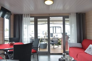 A room with a red sofa, a dining table with chairs, a TV, and a view of boats through large glass doors. - Time out on the Havel - Houseboat "nautikhus" (Werder (Havel))