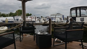 A covered deck with chairs and a table, a boat docked in the background, and a cloudy sky. - Time out on the Havel - Houseboat "nautikhus" (Werder (Havel))