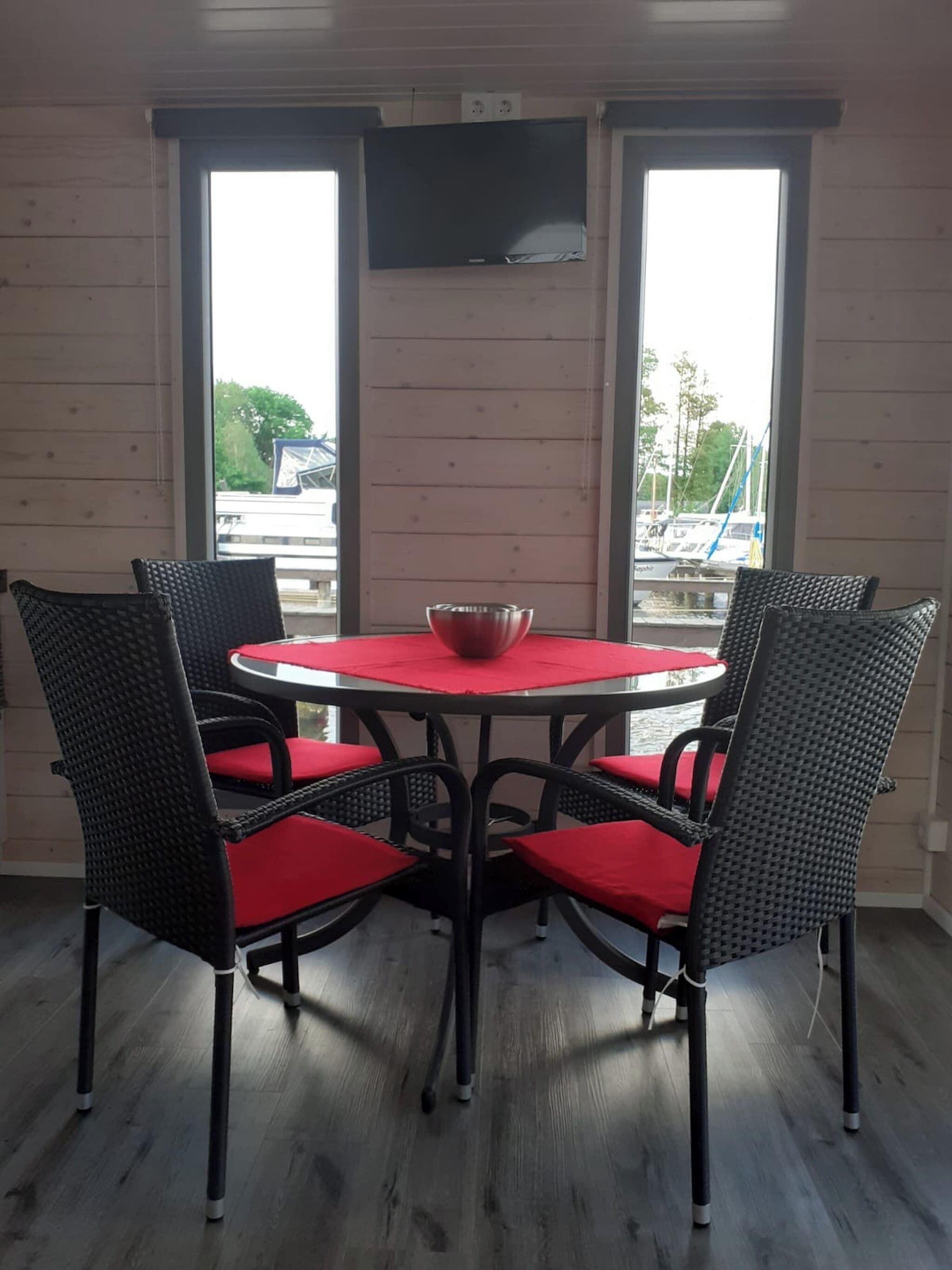 A room with a round table, four chairs, a red cushion, a bowl, and a TV on the wall.