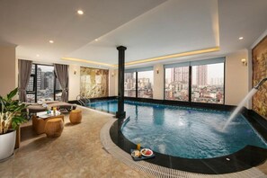 Una piscina infinita en la azotea con vistas a la ciudad, una tumbona y un área de comedor con mesa para dos.
