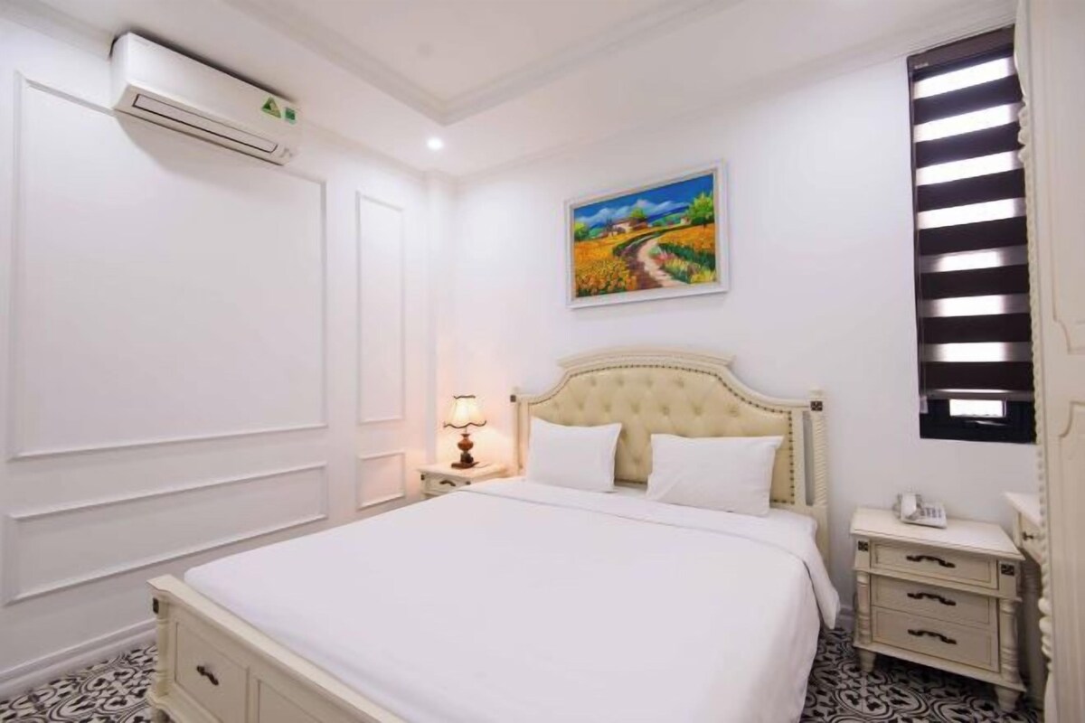 A hotel room with a large bed, a nightstand with a lamp, a painting on the wall, and an air conditioning unit.