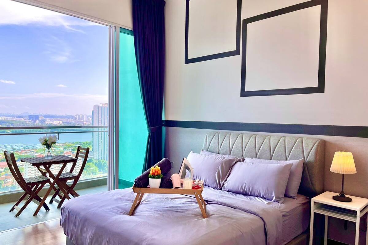 Habitación de hotel con cama, mesita de noche con lámpara, bandeja con artículos de aseo, ventana con vista a la ciudad y balcón con mesa y sillas.