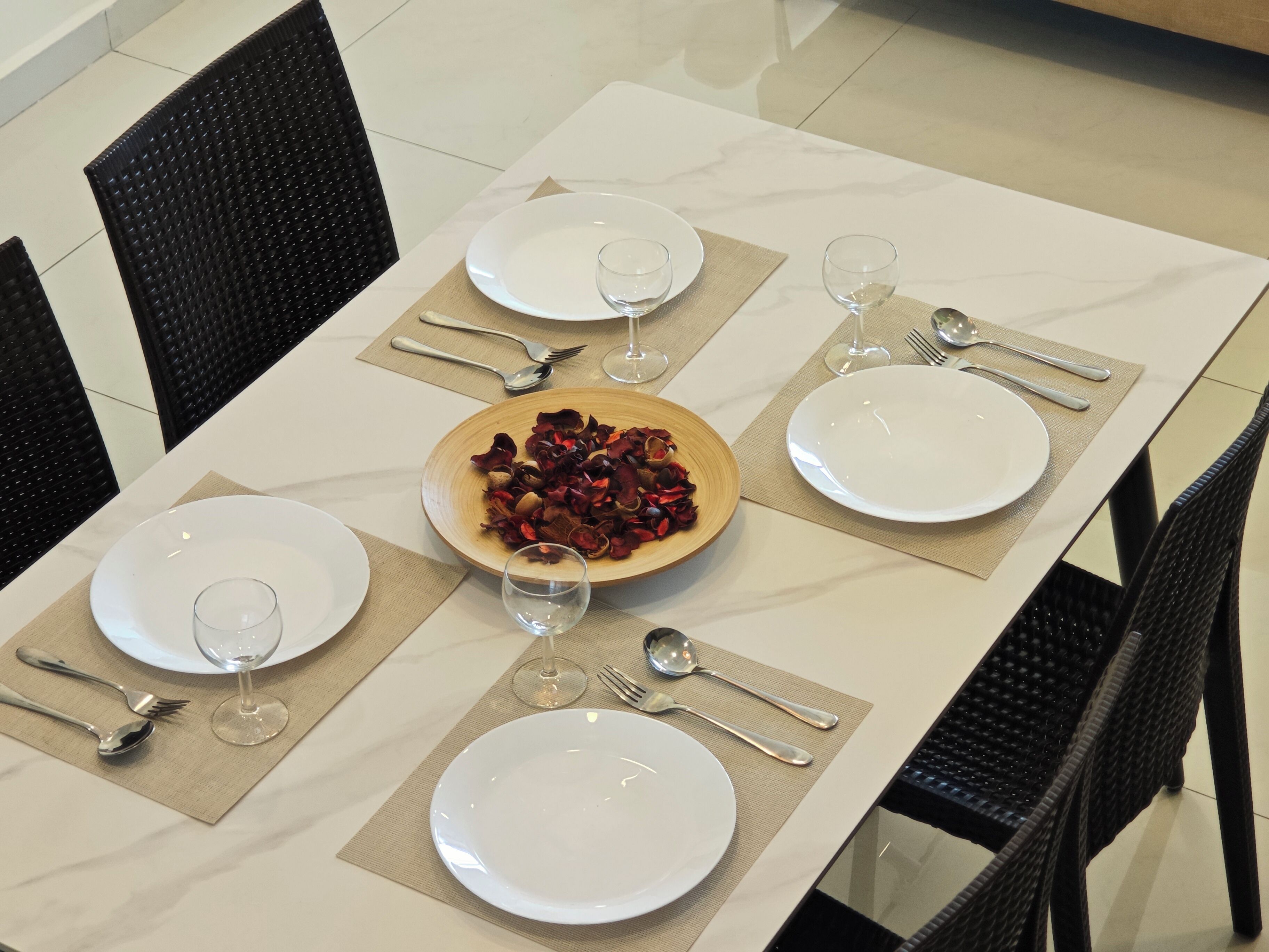 A dining table set with white plates, silverware, and wine glasses, with a bowl of dried rose petals in the center.