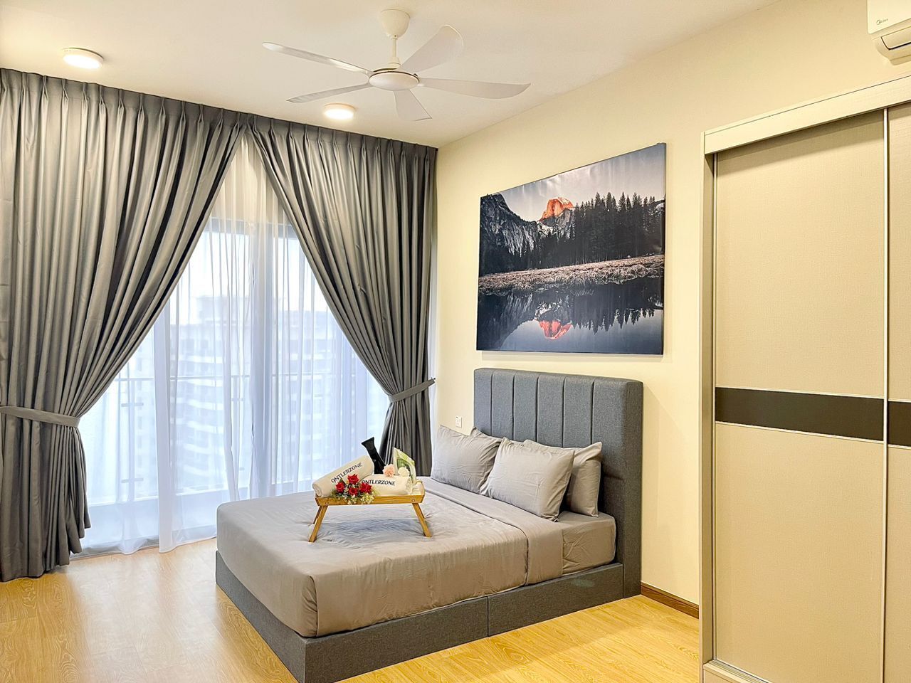 A modern bedroom with a bed, a ceiling fan, a large window with curtains, a painting, and a wooden floor.