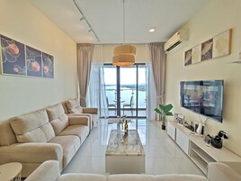 A modern living room with a sofa, a coffee table, and a TV. There is a balcony with a view of the sea.