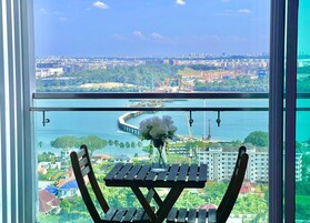 A balcony with a wooden table and chairs, a vase of white flowers, and a cityscape view with buildings and a waterfront.