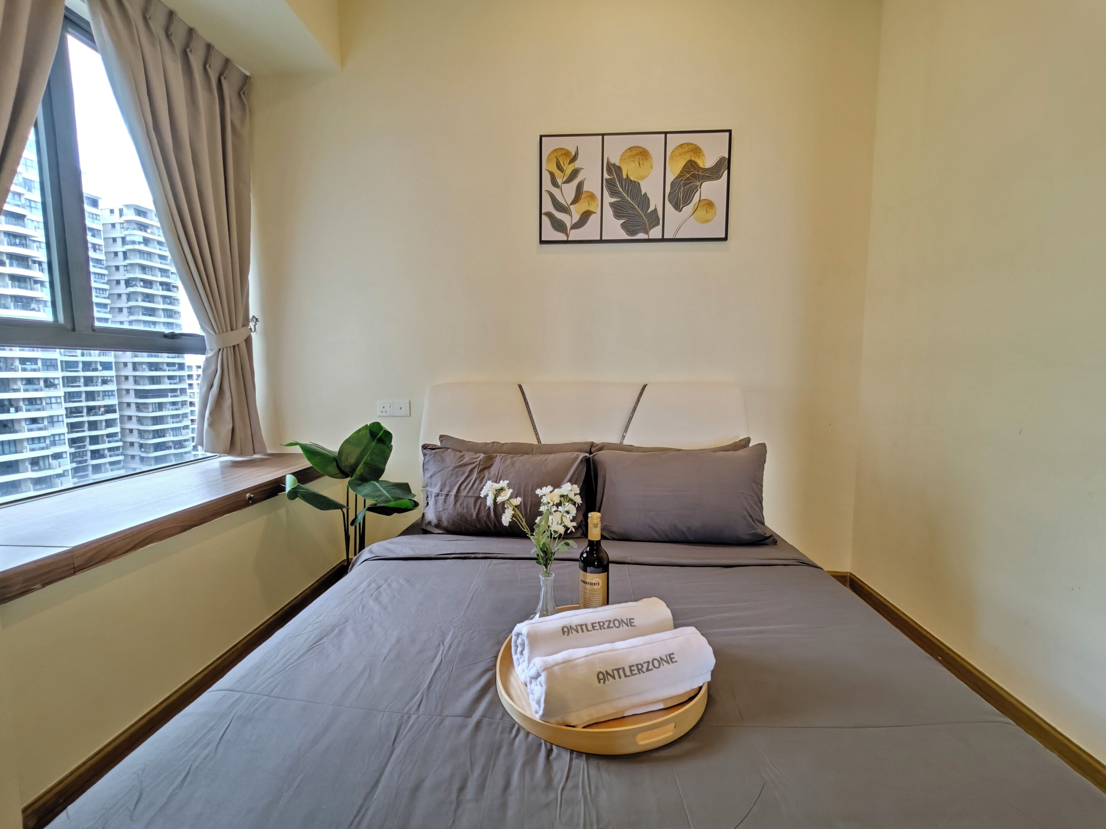Habitació d'hotel amb un llit, una finestra amb cortines, una planta, un cistell amb tovalloles i una ampolla, i una obra d'art a la paret.