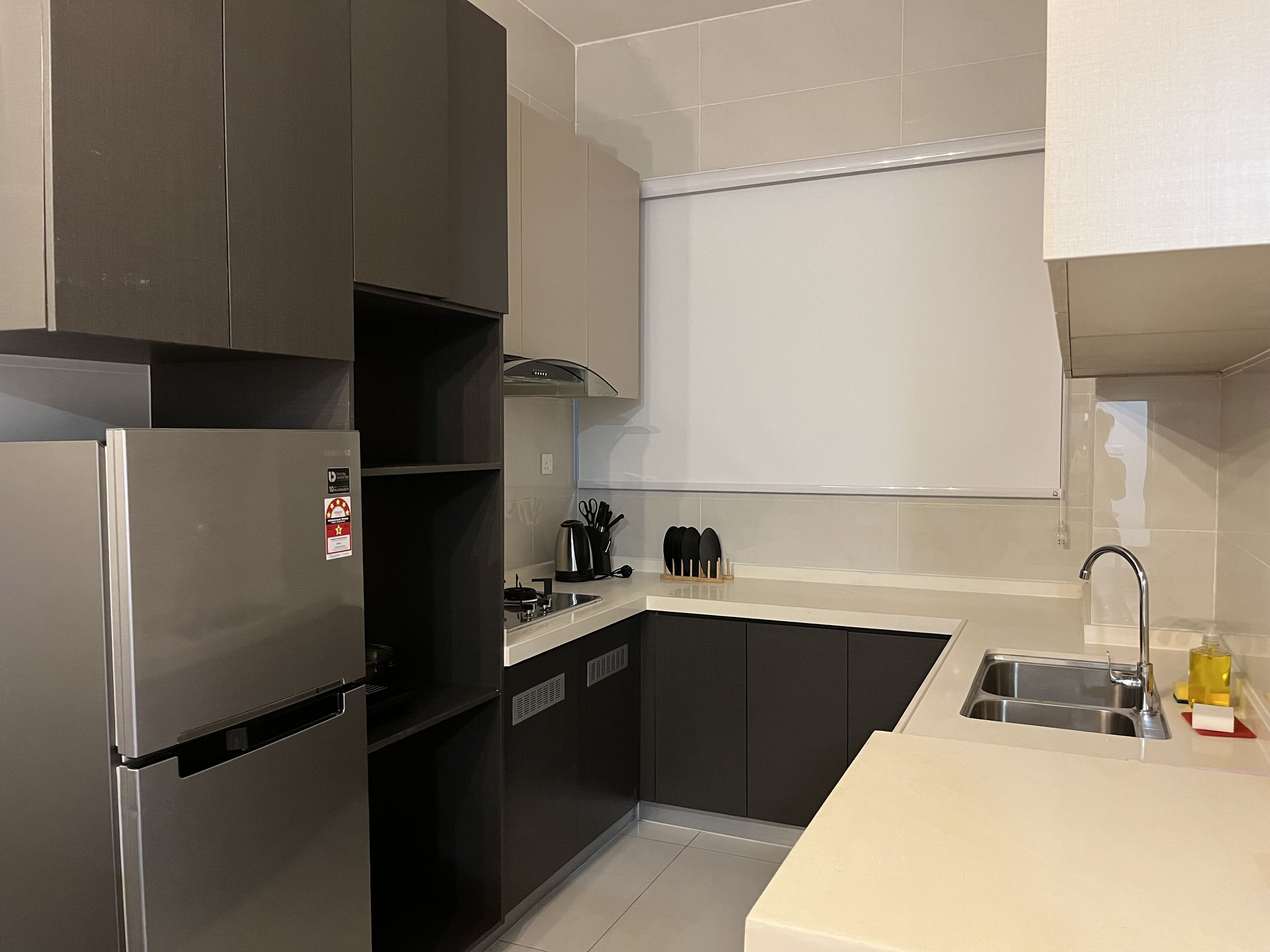 A modern kitchen with dark cabinets, a stainless steel refrigerator, a sink, and a countertop with a kettle and utensils.