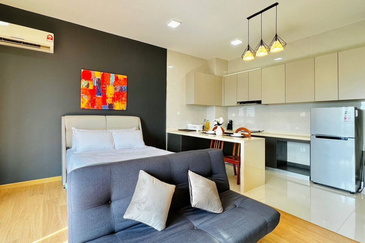 Una habitación de hotel moderna con un sofá gris, una cama con ropa blanca, una zona de cocina con armarios blancos y un cuadro abstracto colorido en la pared.