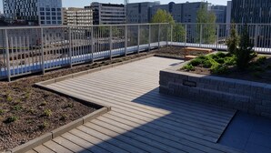 Un jardin sur le toit, doté d’une terrasse en bois, entouré d’une clôture métallique, et offrant une vue sur un paysage urbain avec des bâtiments modernes.