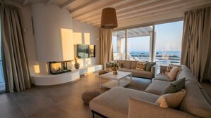 Una sala de estar moderna con un sofá grande, una mesa de centro y un televisor. Se ve el océano a través de amplias ventanas.