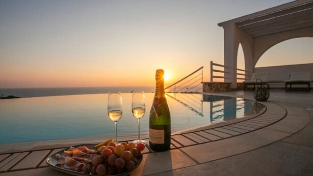 Vista del atardecer desde una piscina infinita, con una botella de vino y copas sobre una bandeja de frutas.
