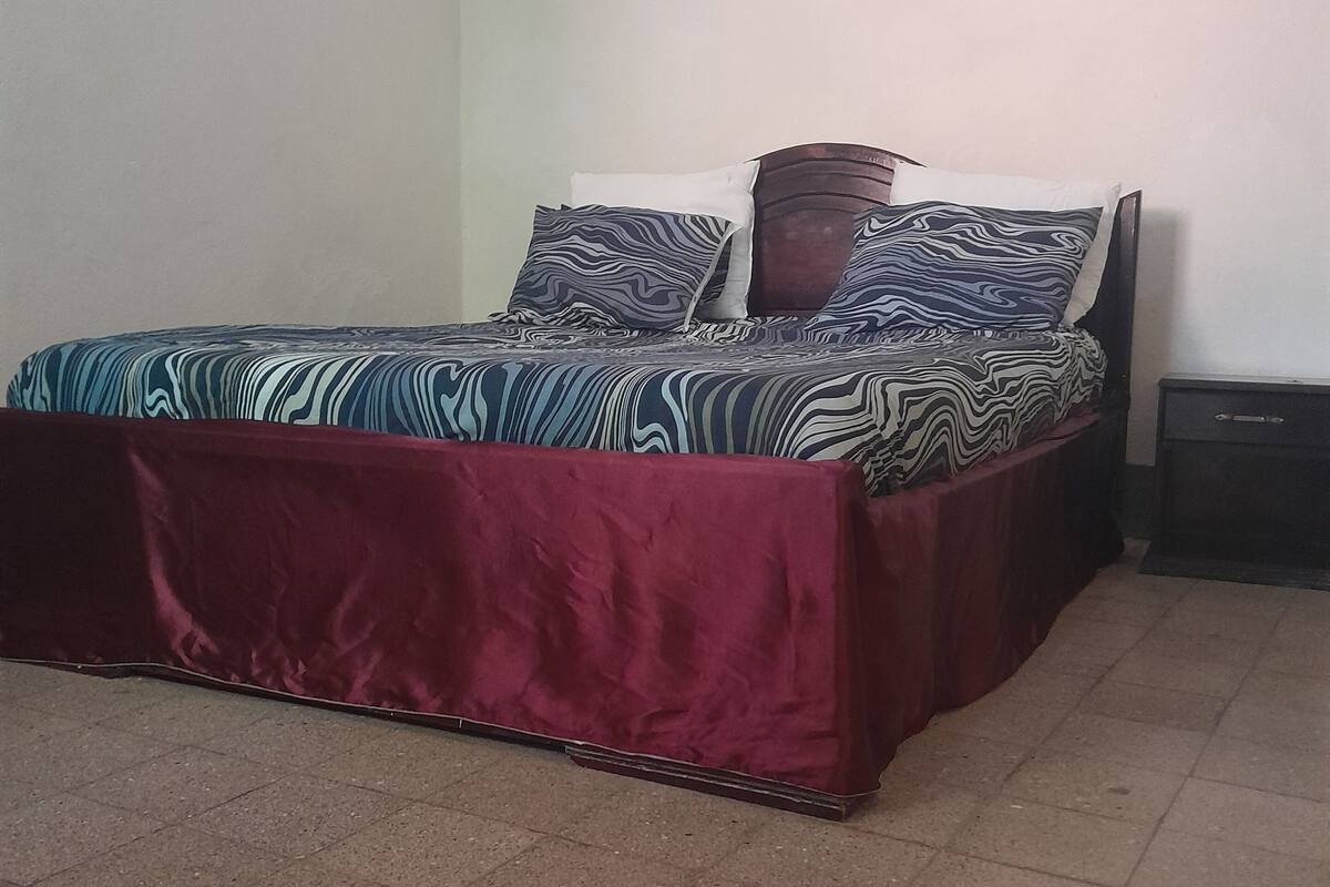 A bed with a patterned bedspread and pillows, a wooden headboard, and a nightstand with a drawer.