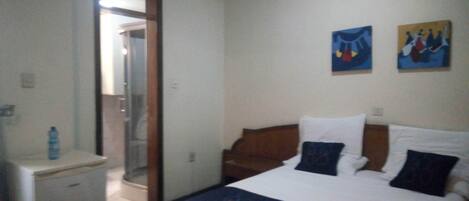 A hotel room with a bed, a small desk, a mirror, and a door leading to a bathroom. There are two paintings on the wall.