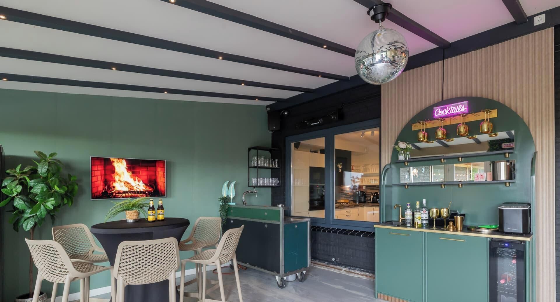 A bar area with a neon sign, a television displaying a fire, and a kitchen visible through the window.