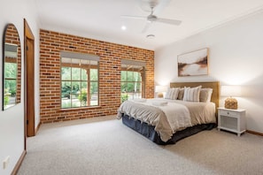 A bedroom with a brick wall, a large window, a bed, a nightstand, a lamp, and a ceiling fan.