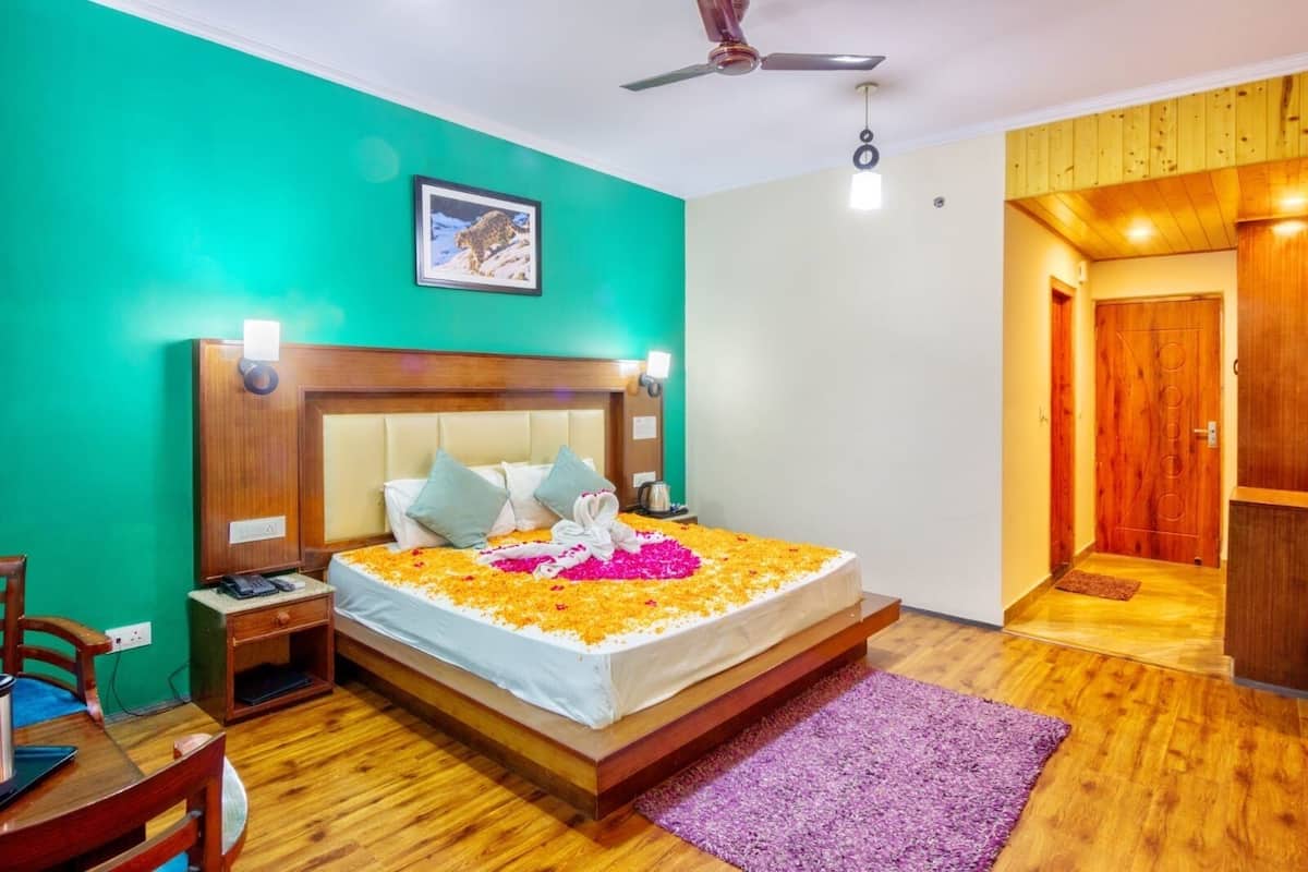 A hotel room with a bed adorned with flower petals, a wooden headboard, a nightstand, a desk with a chair, and a ceiling fan.