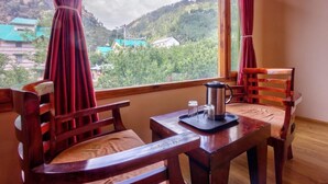 A room with a wooden table and two chairs, a window with curtains, and a view of a mountainous landscape. - Hotel Grand Oasis (Manali)