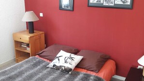 A bedroom with a red wall, a bed with a grey blanket and two pillows, a wooden nightstand with a lamp, and framed pictures on the wall.