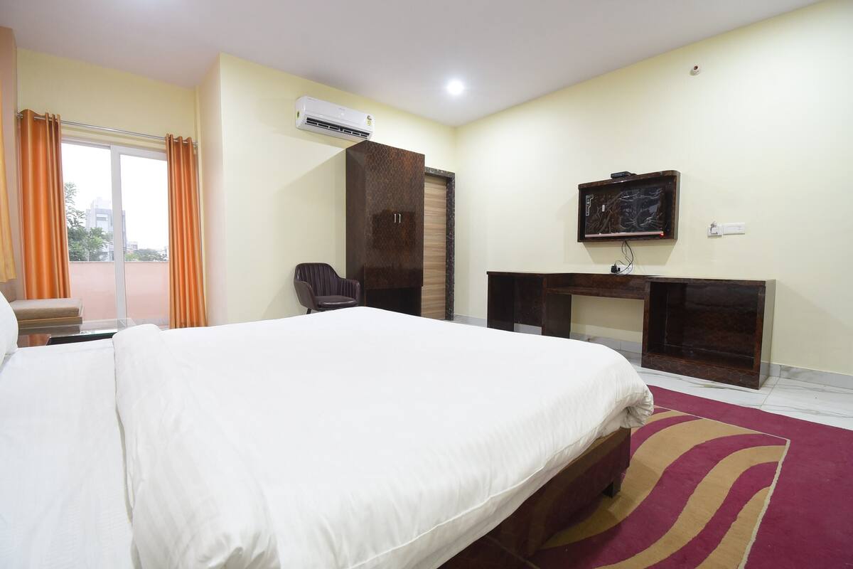 A hotel room with a large bed, a desk, a chair, an air conditioning unit, and a window with curtains.