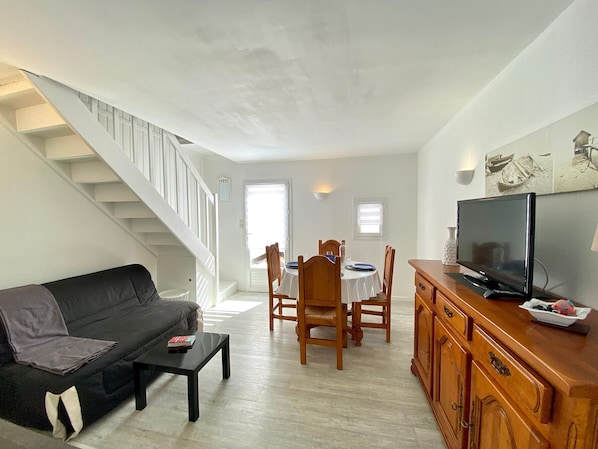 Ein Wohnzimmer mit Sofa, Couchtisch, Fernseher, Esstisch aus Holz mit Stühlen und einer Treppe.