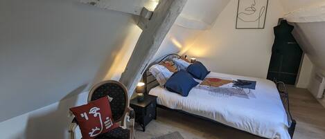 A bedroom with a sloped ceiling, a bed with patterned bedding, a chair with a red pillow, and a wall-mounted artwork.