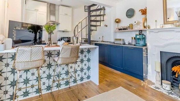 A modern kitchen with blue cabinets, a white island with two chairs, a spiral staircase, and a fireplace. - Beautiful Cottage in Central Whitstable (Whitstable)