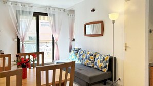 A room with a dining table, chairs, a sofa with patterned cushions, a floor lamp, and a window with sheer curtains.